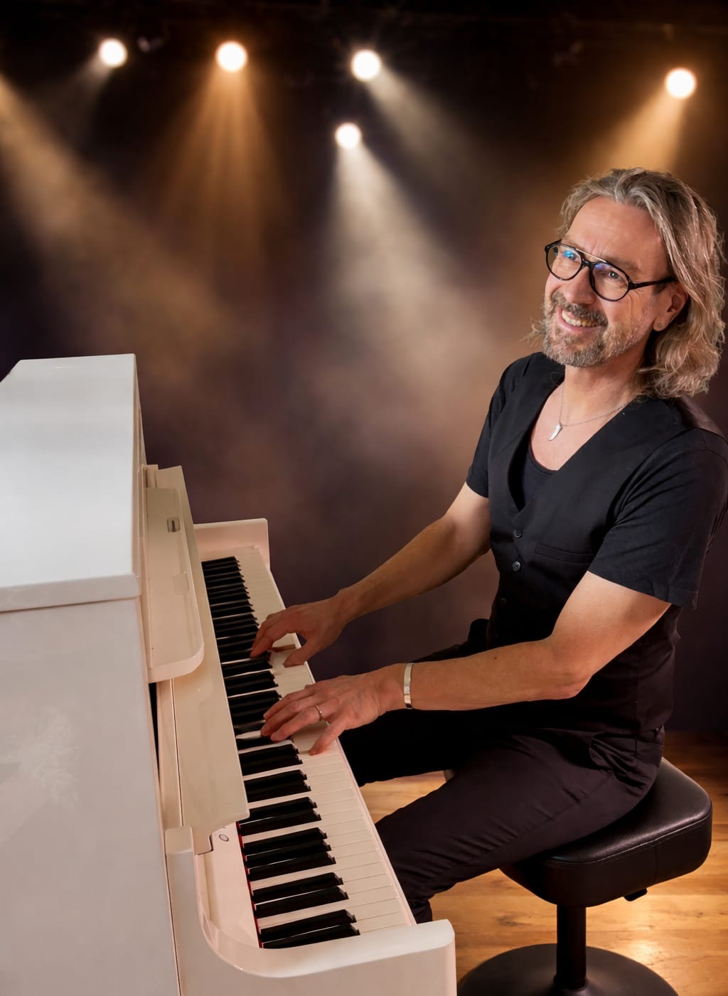 Ein Mann sitzt an einem weißen Klavier. Aus dem Hintergrund bestrahlen ihn Scheinwerfer durch Kunstnebel.